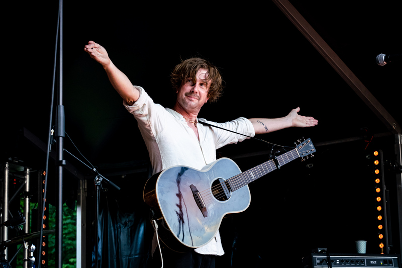 Man med en gitarr står på en scen och sträcker ut armarna i en välkomnande gest
