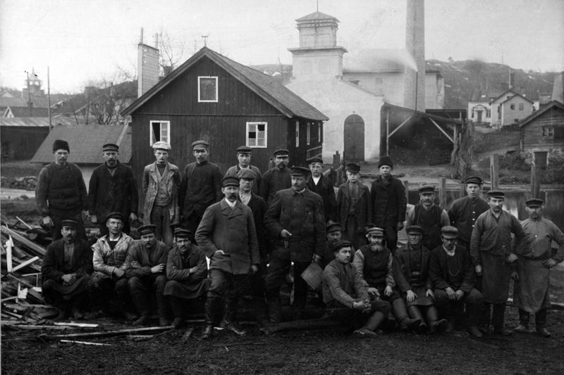 Arbetarna vid Sågbacken. Tjugofem män syns på bilden. Hälften står upp på raden bakom, hälften sitter ner framför. I bakgrunden syns ett industrilandskap med några hus och industribyggnader.