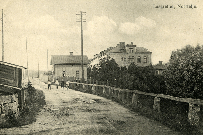 Historisk bild som visar lasarettet i Norrtälje. En grusväg leder dit och fyra pojkar i 10 års åldern går på vägen. Lasarettet är en stor byggnad som sticker upp ovanför träden.