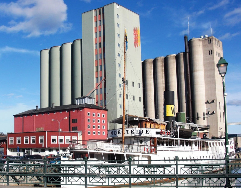 Spannmålssilos i Norrtälje hamn. I förgrunden syns s/s Norrtelje och bakom båten syns en parkering och två röda byggnader. Bredvid dem tornar silosarna upp sig. Den ena är grönmålad med sädeslag målade på framsidan. Den andra silon är grå.
