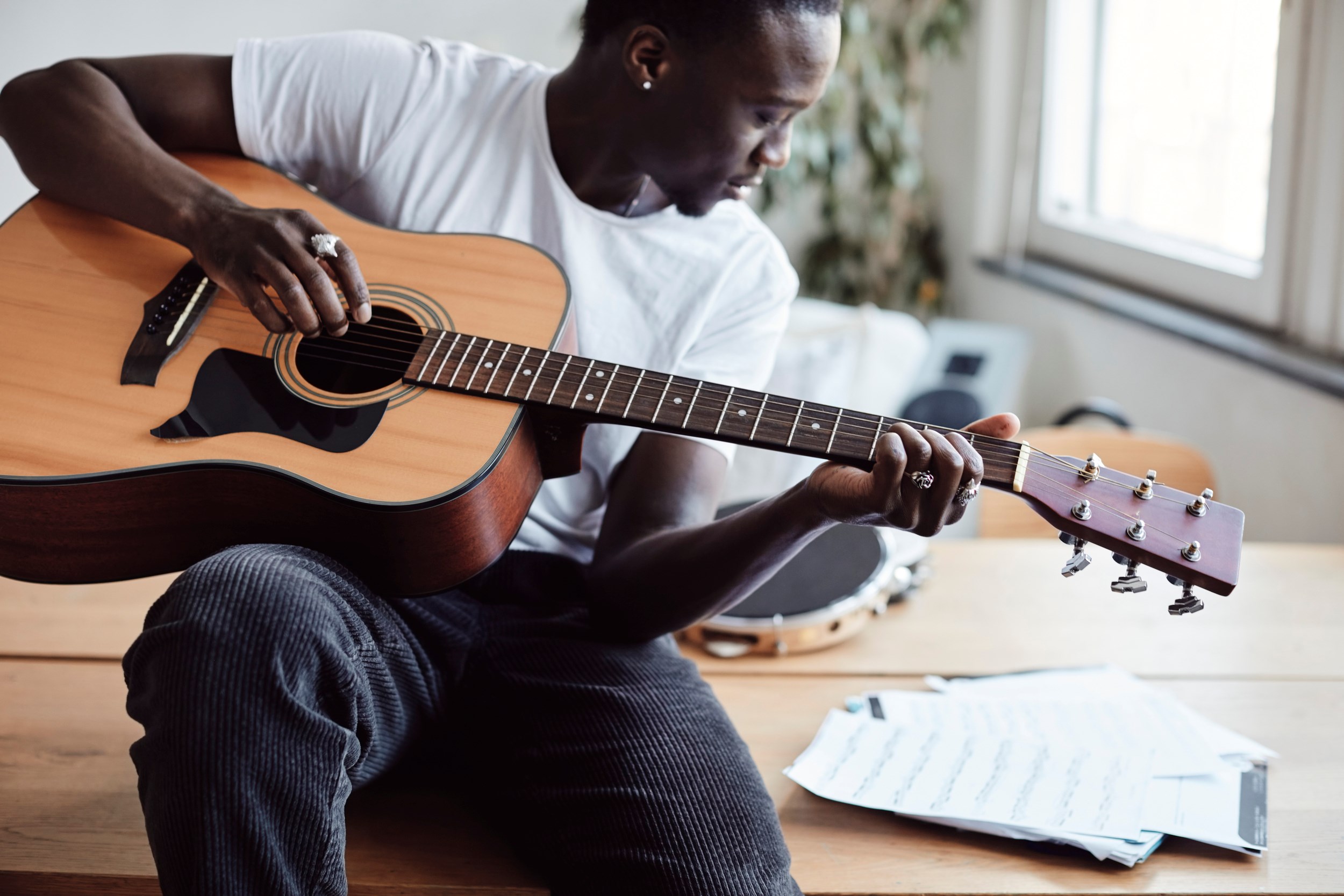 En kille i tonåren sitter på ett bord och spelar gitarr. Han har mörkt hår, vit tröja och svarta byxor. På bordet ligger det papper med noter. 