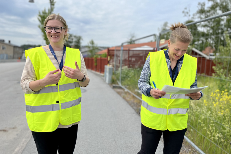 Två kvinnor i reflexvästar står utomhus utanför ett byggstaket och ler.