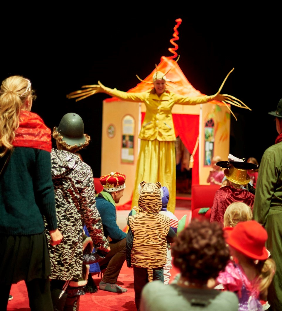 En pedagog utklädd till solen breder ut sina strålar framför en koja. Barn och vuxna i utklädningskläder tittar på.