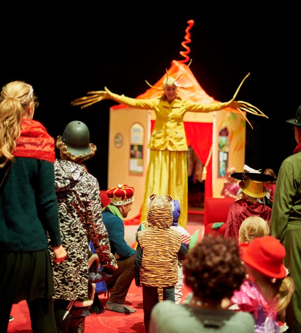 En pedagog utklädd till solen breder ut sina strålar framför en koja. Barn och vuxna i utklädningskläder tittar på.