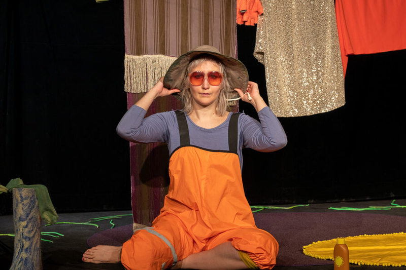 En tjej i orangea galonbyxor sitter på en scen. Hon har blont hår, solglasögon och en regnhatt