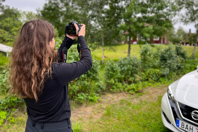 Isabelle Sahba, en kvinna i trettioårsåldern med mörka kläder och mörkt, lockigt hår står med en kamera framför en vit bil och fotograferar ett rött hus.