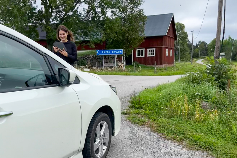 Isabelle Sahba, en kvinna i trettioårsåldern med mörka kläder och mörkt, långt, lockigt hår står med en kamera framför en vit bil.