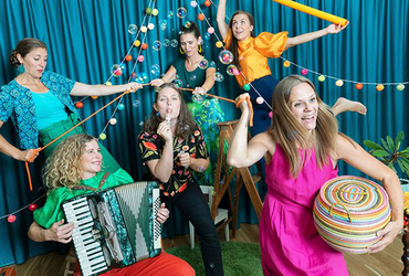 Bilden visar sex kvinnor i ett band som spelar olika instrument och blåser såpbubblor