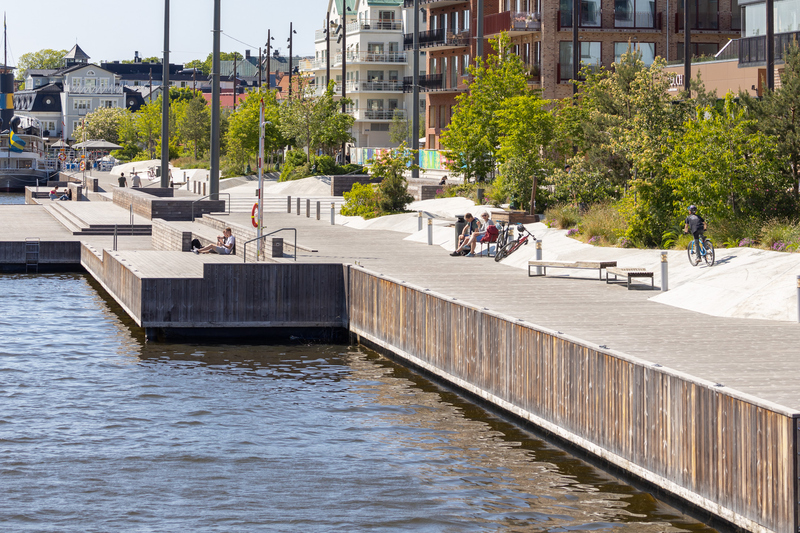 Promenadstråk längs med vattnet en solig junidag. människor som solar, sitter på bänkar, cyklar och promenerar med hund