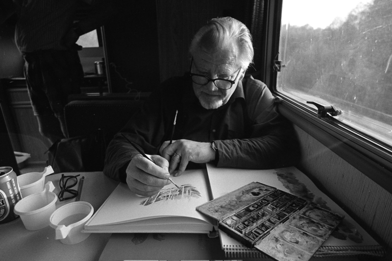 Svartvit bild med en herre som sitter och tecknar i en bok med vita sidor. På skrivbordet står även kaffekoppar. Mannen har vitt hår och glasögon.Fotograf: Roine Karlsson