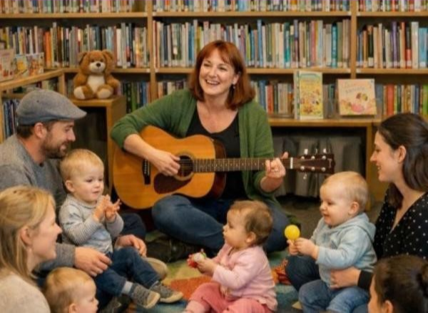 En grupp med föräldrar och små barn sitter i ring kring en vuxen kvinna som spelar gitarr. De sjunger och är glada.