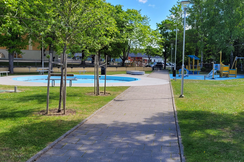 Bilden visar Plaskdammsparken i Hallstavik. På bilden ser man planteringar med perenner och parkbänkar där man kan vila sig i skuggan av ett träd. I parken finns även en plaskdamm och en lekpark.