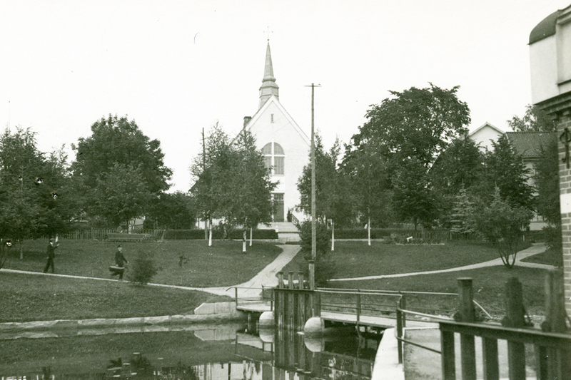 Historisk bild över Norrtälje ån. I förgrunden syns en liten bro över ån, i bakgrunden tornar en vit kyrkoliknande byggnad upp sig.