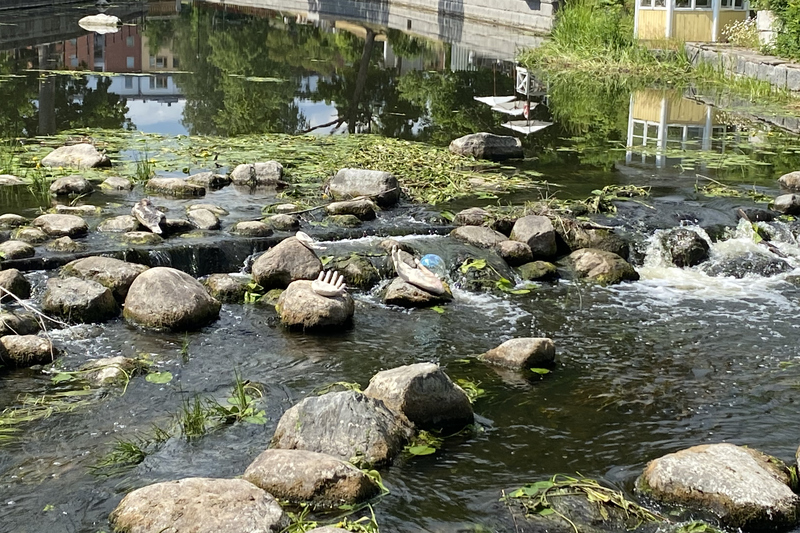 En rinnande å med massor av stenar. I mitten ligger två skulpturer som är två händer och den ena handen håller i en genomskinlig pärla.