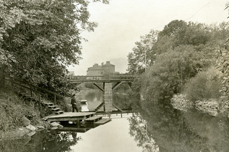 Landskapsbild. Norrtäljeån med grönska runt om och en bro längre bort i bilden