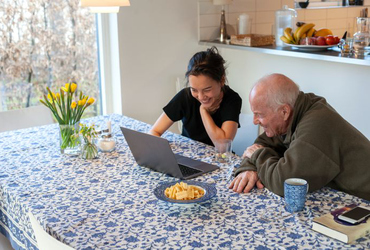 Äldre man och yngre kvinna tittar pĺ en laptop vid ett köksbord.