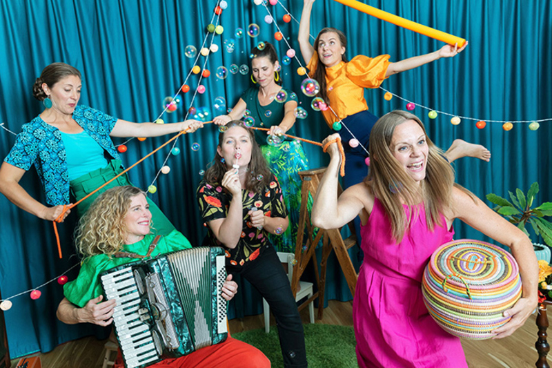 En band med sex kvinnor syns på bilden. En spelar trumma, en dragspel och en blåser såpbubblor