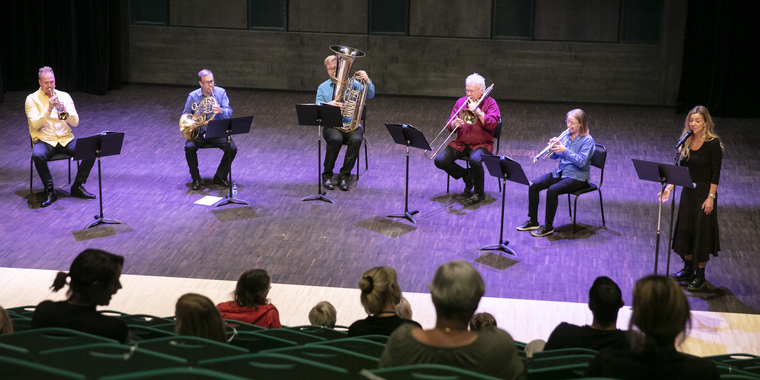 Bild tagen på Rimbo kulturscens scen från läktaren. På scenen sitter spelar fem personer instrument och en person sjunger i mikrofon.