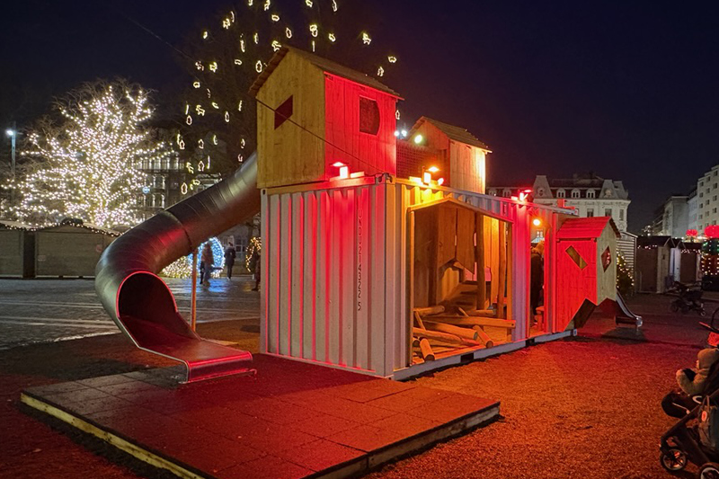 Ett torg i skymning, det är mörkt förutom julbelysning som lyser upp platsen och en vit container med en lekstuga på taket och en rutchkana på ena sidan som lyser.