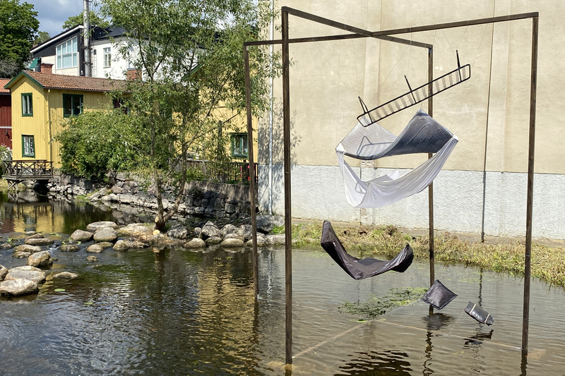 I Norrtäljeån står en stor ram. I ramen hänger en säng i flera delar. Högst upp stommen, sen madrass, lakan, täcke och längst ner mot vattnet, två kuddar.