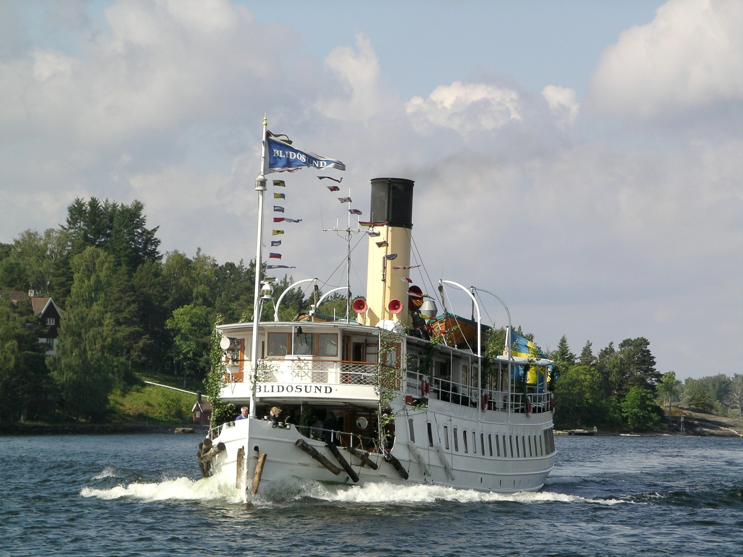 En bild på ångbåten Blidösund som åker med fart i det blåa vatten. I bakgrunden syns gröna träd, moln och ett rött hus som gömmer sig bakom träden.