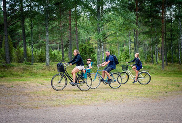 En familj cyklar pÃ¥ rad pÃ¥ grusvÃ¤g fÃ¶rbi kameran