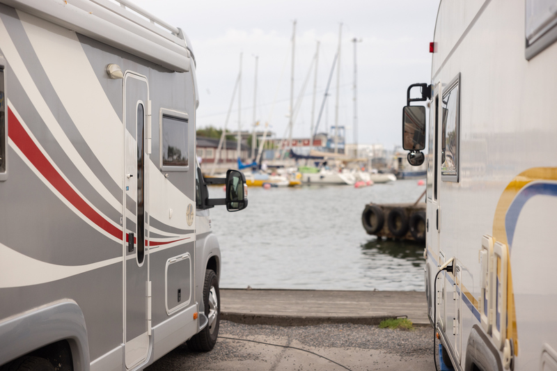 två husbilar står parkerade med fronten mot en kaj. I bakgrunden syns båtar och en brygga.