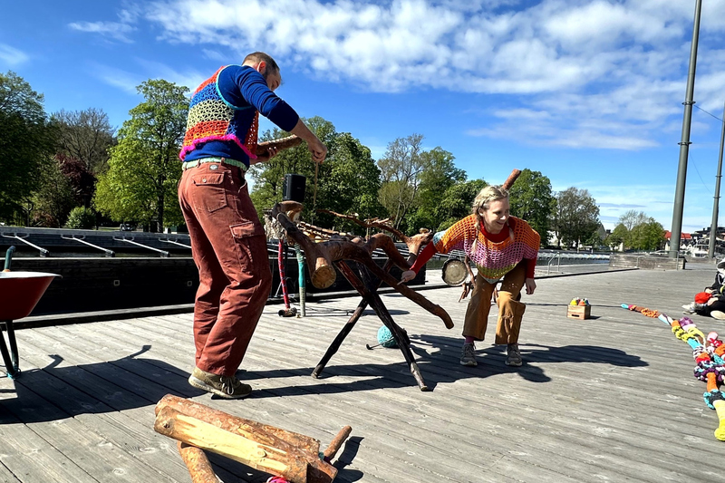 En man och en kvinna i färgglada virkade kläder dansar och trummar på en trädgren