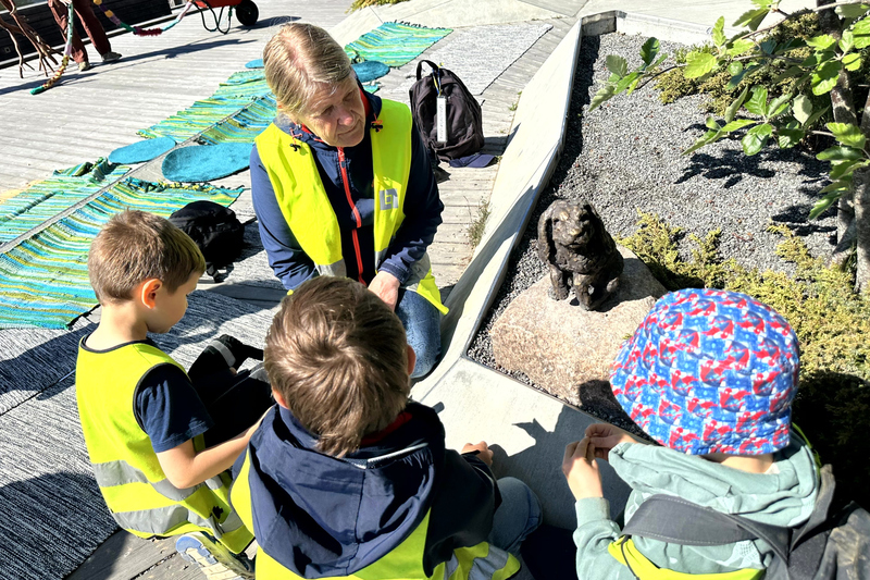 tre barn och en pedagog i reflexvästar sitter runt en kanin i brons och pratar