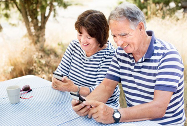 Ett pensionärspar sitter utomhus vid ett bord och tittar på sina mobiler