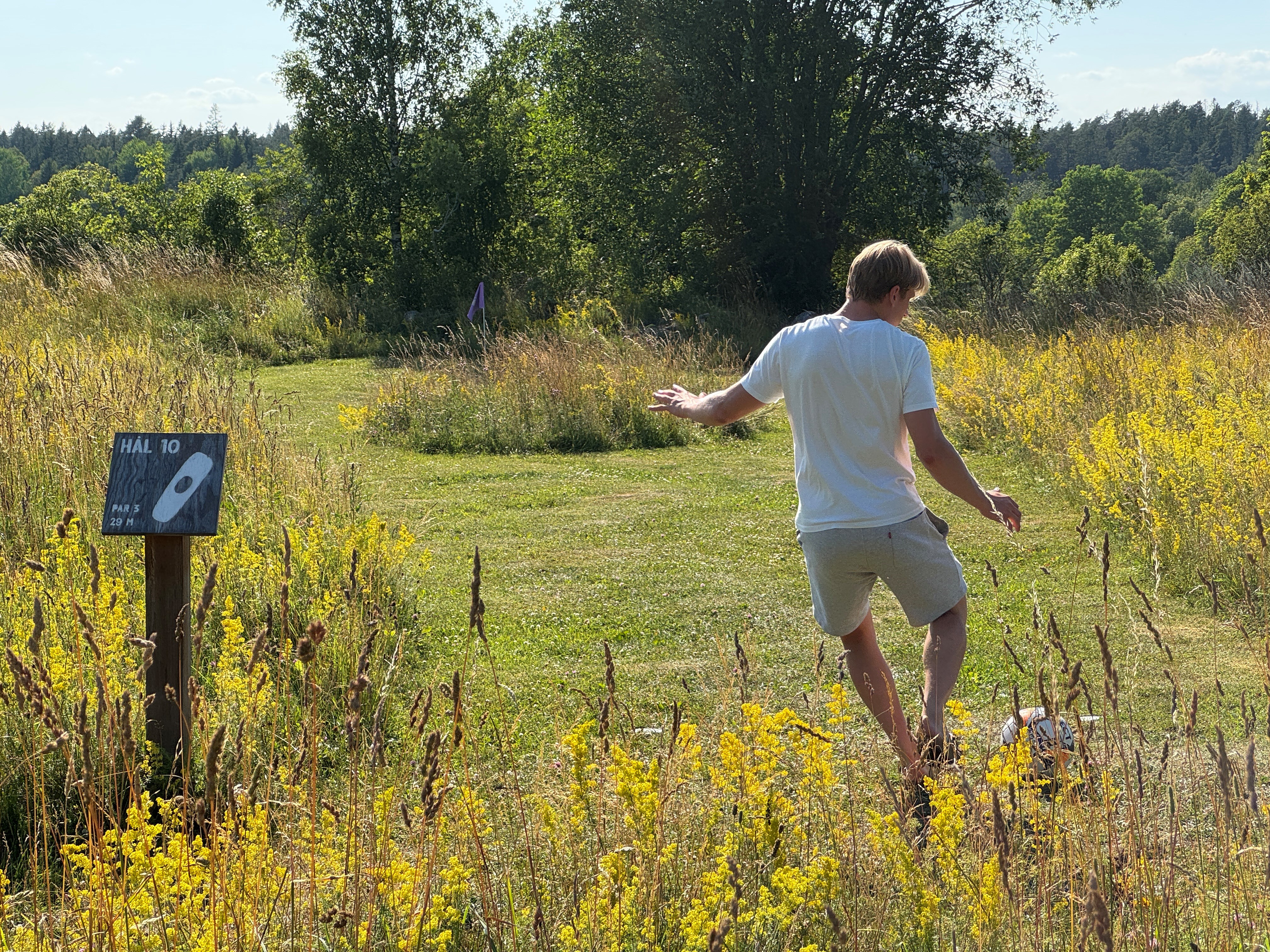 En person spelar fotbollsgolf på en grön bana omgiven av sommargräs, blommor och natur vid Norrtälje Fotbollsgolf.