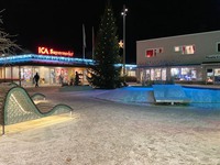 Bild som föreställer ett upplyst torg. En stor julgran står i centrerad i bild  och vid sidan av julgranen ses en person gå in genom dörrarna på en ICA Supermarket-butik.