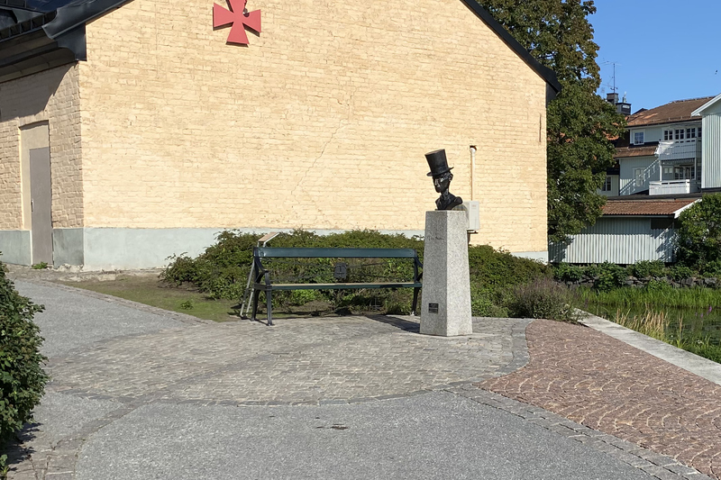 Bilden visar en vy över Nils Ferlins gränd i Norrtälje stad. På bilden ser man en parkbänk och en statyett över Nils Ferlin.